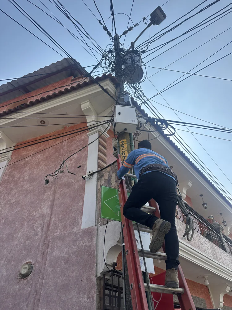 Técnico subido a una escalera trabajando junto a un gabinete instalado en un poste con cableado aéreo, frente a la fachada de un edificio.
