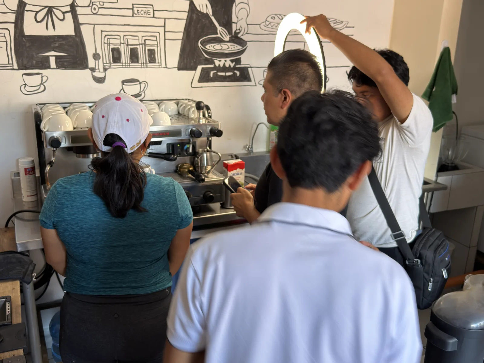Equipo de NT502 grabando dentro de una cafetería: una persona sostiene un aro de luz encendido mientras otra prepara la toma con un celular junto a la máquina de espresso, donde trabaja una barista; en la pared, un mural en blanco y negro de una cocina.
