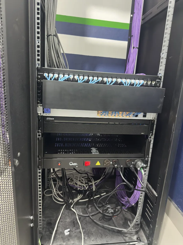 Rack de comunicaciones abierto con un patch panel de 24 puertos y cables de parcheo azules, un organizador horizontal, un switch, un grabador de video y una regleta de energía; por los lados bajan haces de cable gris y morado.