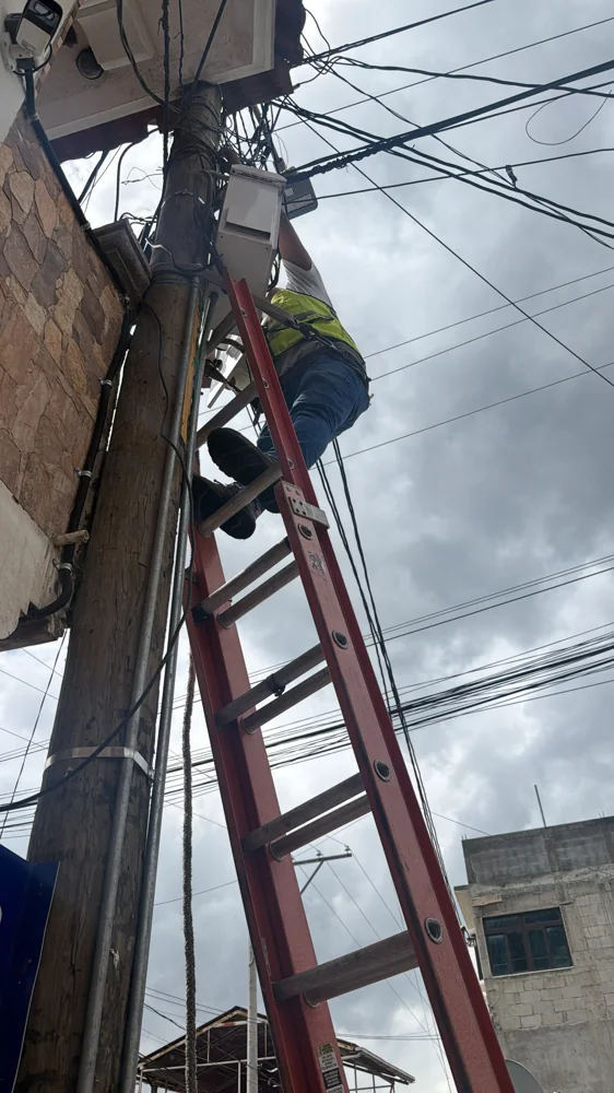 Técnico con chaleco reflectivo en lo alto de una escalera, instalando equipo en un poste de madera junto a un muro de piedra.
