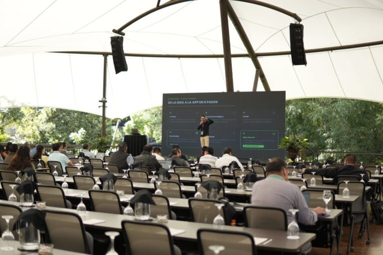 Conferencista con micrófono frente a una pantalla con la presentación «De la idea a la app en 6 pasos», ante el público bajo una carpa.