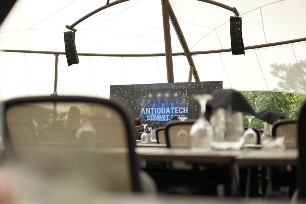 Pantalla con el logo «Antigua Tech Summit 2026» vista entre sillas y copas del salón.